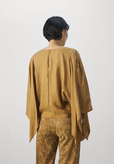 Blusa di seta color senape con maniche larghe e fluenti e zip sul retro. Abbinata a pantaloni con motivo floreale testurizzato.