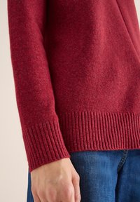 Maglione rosso lavorato a maglia con polsini e orlo a coste. La texture è morbida con un sottile motivo mélange, abbinato a jeans blu.