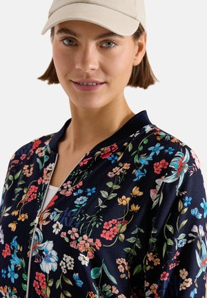 Ung kvinde iført beige kasket og marineblå blomstret lynlåstrøje over hvid top, smilende let, med kort brunt hår synligt.