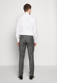 Weißes Langarmhemd, slim fit, kombiniert mit grauen maßgeschneiderten Hosen mit zwei Gesäßtaschen und schwarzen Knopfakzenten, getragen mit schwarzen Schuhen.