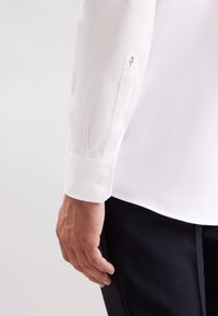 Camicia a maniche lunghe bianca a chevron con polsini a bottone, caratterizzata da un delicato dettaglio ricamato sulla manica. Tessuto morbido e leggero.