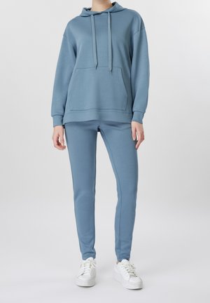 Personne portant un sweat à capuche bleu et un pantalon de jogging assorti avec des baskets blanches, debout devant un fond clair uni.