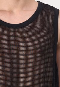 Close-up van een persoon die een mouwloos zwart mesh-shirt draagt, waarbij de huid en borst onder de rasterpatroonstof zichtbaar zijn.
