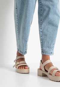 Béžové platformové sandály s dvěma nastavitelnými popruhy a strukturovanou podrážkou; nošeny s světle modrými ohrnutými džínami.