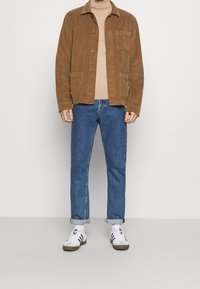 Veste en velours côtelé marron avec deux poches avant, col roulé beige, jean en denim bleu et baskets blanches avec des accents marron.