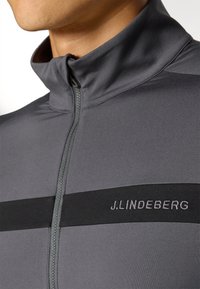 Gray zip-up top featuring a high collar, solid black horizontal stripe, and embroidered "J.LINDEBERG" logo on the chest. Smooth fabric texture.