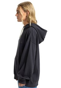 Zwarte oversized hoodie met een buidelzak, witte trekkoorden en logo-detail op de borst. Zacht materiaal, casual pasvorm.