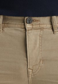 Los pantalones de algodón color beige presentan un diseño clásico de cinco bolsillos, un cierre de botón metálico y costuras visibles para un detalle adicional.