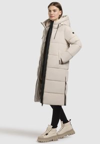 Langer beigefarbener Puffer-Mantel mit Kapuze, ausgestattet mit einem Reißverschluss, elastischen Akzenten und seitlichen Taschen. Kombiniert mit beigen klobigen Schnürstiefeln.