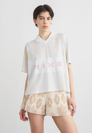 Personne portant un polo blanc Nike avec un logo rouge et une jupe plissée beige à motif floral sur un fond blanc uni.