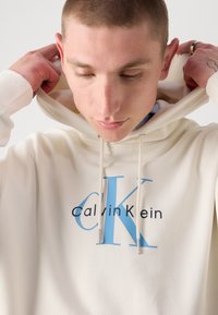 Sudadera color crema hecha de una tela suave. Presenta un gran logo "CK" en azul y texto en negro. Cuerdas de ajuste con acentos plateados.