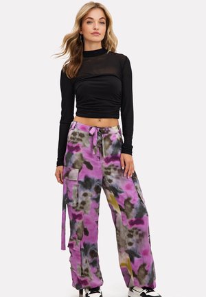 Vrouw die een zwart doorschijnend cropped top met lange mouwen draagt en losse paarse, grijze en groene tie-dye cargobroek met sneakers, staand tegen een witte achtergrond.