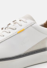 Weiße Ledersneaker mit einer glatten Oberseite, grauen Akzenten, einer strukturierten Gummisohle und einem kleinen gelben Etikett in der Nähe der Schnürsenkel.