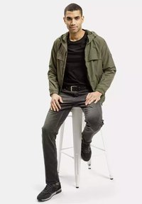 Jasnozielona, lekka kurtka z kapturem, zestawiona z czarną koszulą i ciemnoszarymi dżinsami, noszona z czarnymi trampkami na stołku.