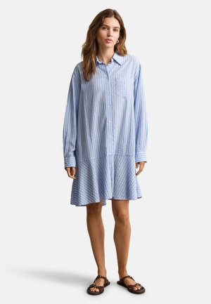 Junge Frau trägt ein lockeres, langärmeliges, blau-weiß gestreiftes Hemdkleid und braune Sandalen, steht vor einem einfarbigen Hintergrund.