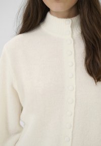 Egy nő fehér gombos kötött pulóvert visel, magas bordázott gallérral, hosszú, barna haja egyik vállára lóg.