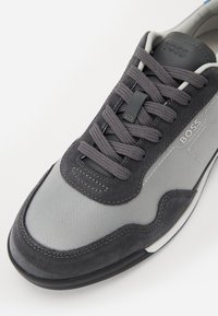 Chaussure de sport grise avec un mélange de daim texturé et de tissu en mesh. Comprend des lacets ronds, une languette rembourrée et une semelle en caoutchouc noire contrastante.