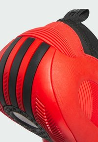 Zapatilla deportiva roja con acentos de goma negra, superficie texturizada y una suela redondeada que presenta surcos distintivos y un detalle central circular.