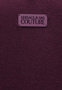 Burgundijas adīta auduma ar teksturētu virsmu, kurā ir metāliski diegi. Pie tā ir piestiprināta violetas krāsas taisnstūra birka ar uzrakstu "VERSACEJEANS COUTURE".