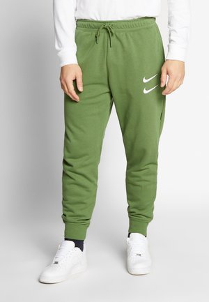 Mann trägt grüne Nike Sweatpants mit doppeltem weißem Swoosh-Logo, schwarze Socken, weiße Sneakers und ein weißes Langarmshirt.