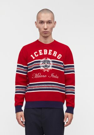 Uomo che indossa un maglione a righe rosse con la scritta e l'emblema "Iceberg Milano Italia", in piedi davanti a uno sfondo chiaro uniforme.