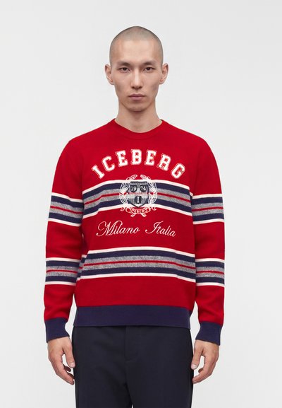 Homme portant un pull rayé rouge avec le texte et l'emblème « Iceberg Milano Italia », debout devant un fond clair uni.