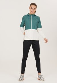 Teal en witte kleurgeblokte lichtgewicht jas met een ritssluiting aan de voorkant en capuchon; gecombineerd met zwarte nauwsluitende broeken en grijze sportschoenen.