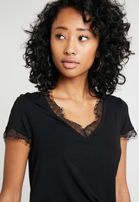 T-shirt noir à manches courtes, avec une bordure en dentelle autour du décolleté en V et des bords de manches. Tissu doux avec une coupe ajustée.