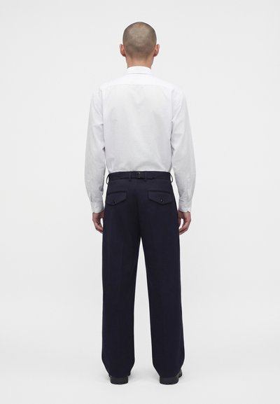 Personne à la tête rasée portant une chemise blanche à manches longues et un pantalon ample foncé, debout de dos sur un fond blanc uni.