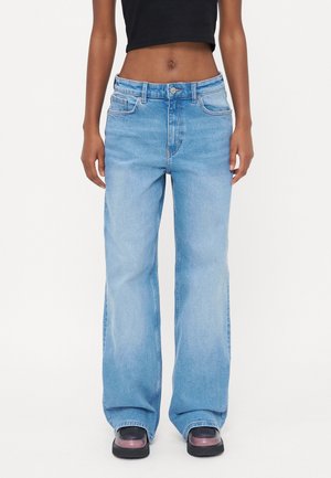 Persoon draagt lichtblauwe wijde jeans gecombineerd met een zwarte crop top en roze en zwarte schoenen, staand tegen een effen achtergrond.