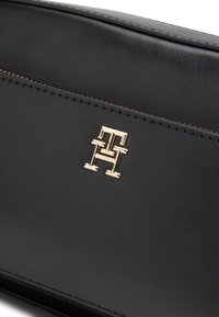 Geantă neagră din piele, cu cusături vizibile și un mic emblemă „TH” aurie, întrețesută, plasată central pe suprafața din față.