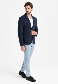 Marineblazer mit schmalen Revers, zwei Vordertaschen, kombiniert mit einem weißen T-Shirt und hellblauen Jeans, ergänzt durch schwarze Loafers.