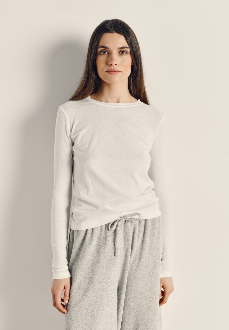 Žena s dugom smeđom kosom, odjevena u bijelu majicu dugih rukava i sive trenirke, stoji nasuprot jednostavnoj svijetloj pozadini.