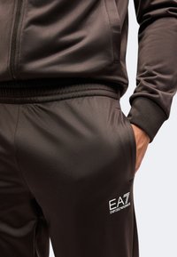 Κοντινή λήψη ενός ατόμου που φοράει σκούρο καφέ φόρμα EA7 Emporio Armani με το χέρι στην τσέπη, εμφανίζοντας το λογότυπο στο παντελόνι.