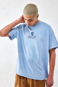 Light blue cotton T-shirt with short sleeves, featuring a graphic design in dark blue text on the chest. Model wears brown corduroy pants.