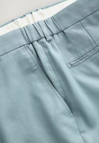 Lichtblauwe geweven broek met een elastische tailleband, achterzak en een gladde textuur. Zichtbare stikseldetails langs de naden.