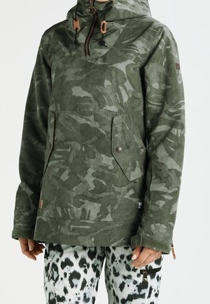 Jacke mit grünem Camouflage-Muster, Kapuze, Zugbändern, Reißverschluss und zwei Fronttaschen, kombiniert mit schwarz-weißen gepunkteten Hosen.