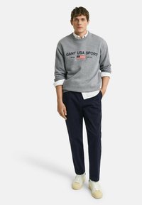 Sweatshirt gris avec le texte "GANT USA SPORT" et le drapeau américain. Associé à un pantalon bleu marine foncé et des baskets blanches avec des accents beiges.