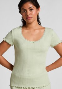 Vrouw in een lichtgroene ribgebreide top met korte mouwen, scalloped randen en een klein strikje bij de halslijn, met haar handen op de heupen.