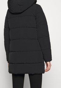 Černá prošívaná péřová bunda s kapucí, nošená osobou otočenou zády, ukazující záda a část černých kalhot.