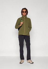 Olivno zelena flis jakna z visokim ovratnikom in zadrgo spredaj. Črne cargo hlače z bočnimi žepi in bež pohodni čevlji. Dodatki vključujejo zelen nahrbtnik in sončna očala.