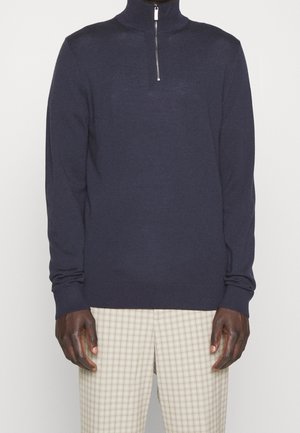 Homme portant un pull à manches longues bleu marine foncé avec une fermeture éclair frontale argentée et un pantalon beige à carreaux, sur un fond uni.