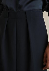 Tumši zils svārku plisēts audums ar stingru struktūru, izceļas ar augstu vidukli un tīrām līnijām. Gluda tekstūra ar vieglu spīdumu.