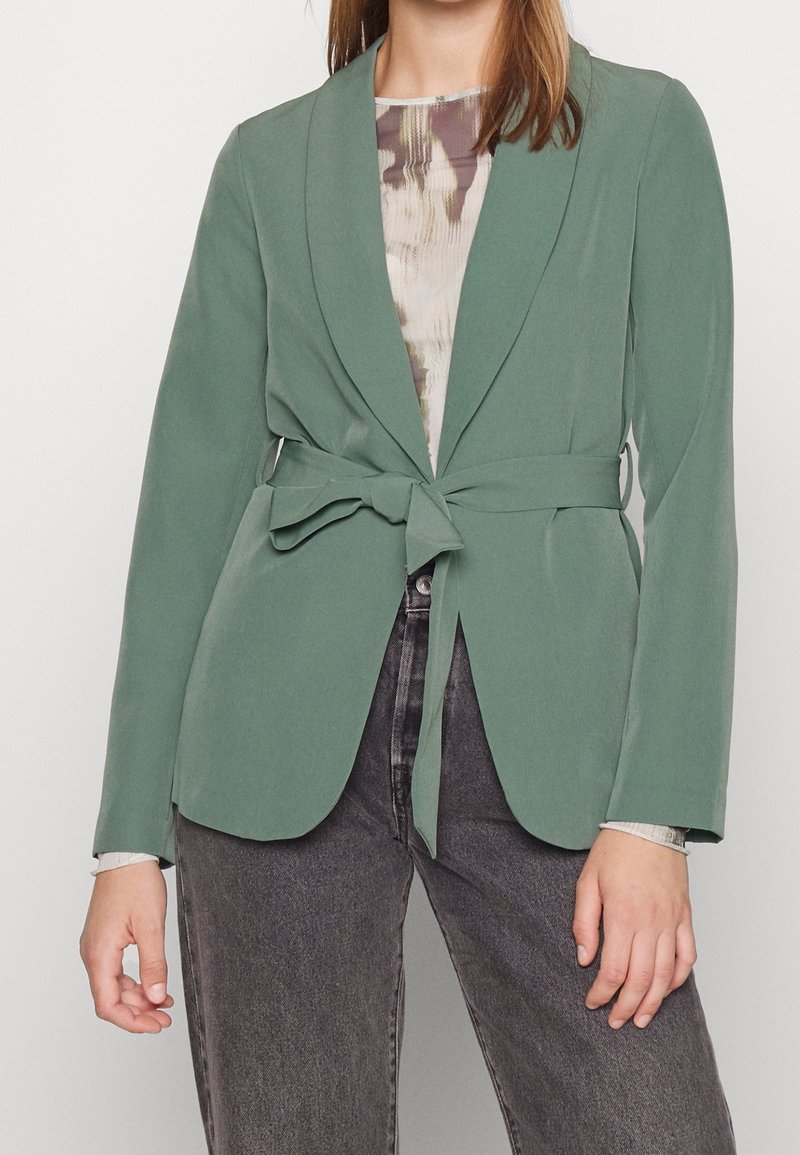 Blazer vert avec une ceinture à nouer, col à revers et manches longues. Associé à un haut à motif et un jean foncé, offrant une texture lisse.