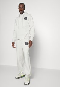 Hvid hættetrøje og matchende sweatpants lavet af blødt stof. Har en frontlomme, logo-patch og kontrasterende syninger. Sneakers med grønne detaljer.