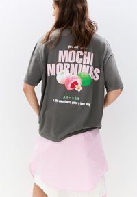 T-shirt gris à manches courtes avec un graphisme coloré de mochi et le texte "MOCHI MORNINGS." Superposé sur une jupe rayée rose clair.