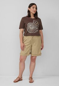 Braunes Baumwoll-T-Shirt mit Spiralgrafik, kombiniert mit khakifarbenen Shorts mit umgeschlagenen Säumen und elastischem Bund. Beige Sandalen runden das Outfit ab.