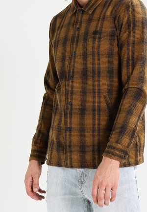 Homme portant une veste à carreaux marron et noir avec des boutons-pression, associée à un jean bleu clair, visible du torse aux mains.