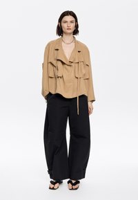 Tan, gelaagd top met een diepe V-hals, voorbindstrikken en meerdere zakken; gecombineerd met losse zwarte broeken en platte sandalen met accenten.
