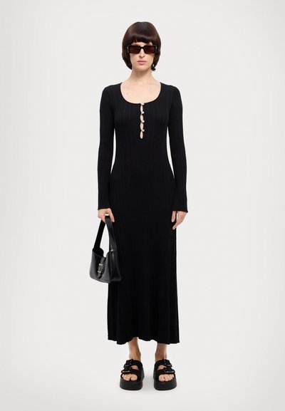 Black ribbed maxi dress with long sleeves and a rounded neckline, featuring button detailing. Worn with platform sandals and a black handbag.